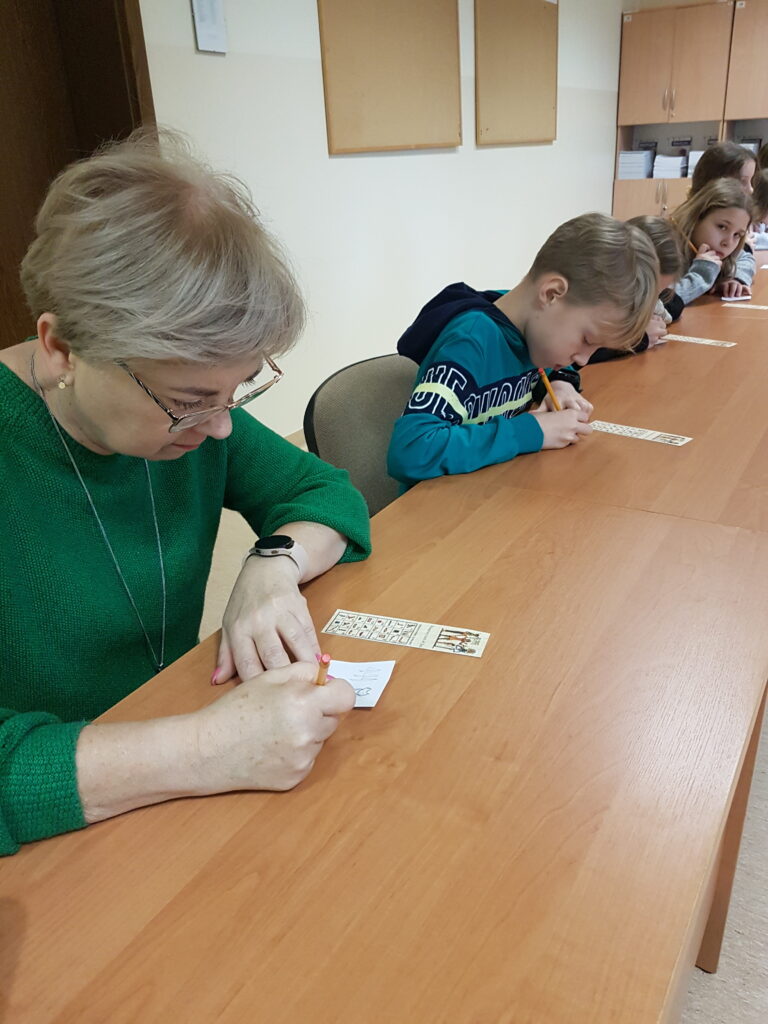 Nauczycielka w okularach i zielonej sukience siedzi przy długim stole. Pisze na małej kartce. Przed nią leży zakładka do książki. W tle przy stole siedzą piszący uczniowie.