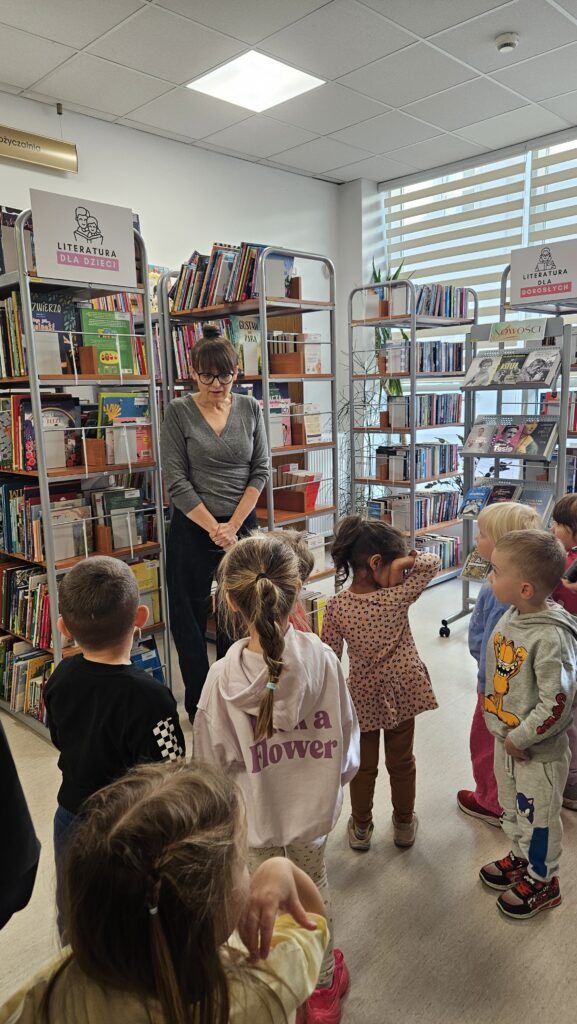 Grupa dzieci w wypożyczalni. Bibliotekarka stoi i opowiada. W tle regały z książkami.