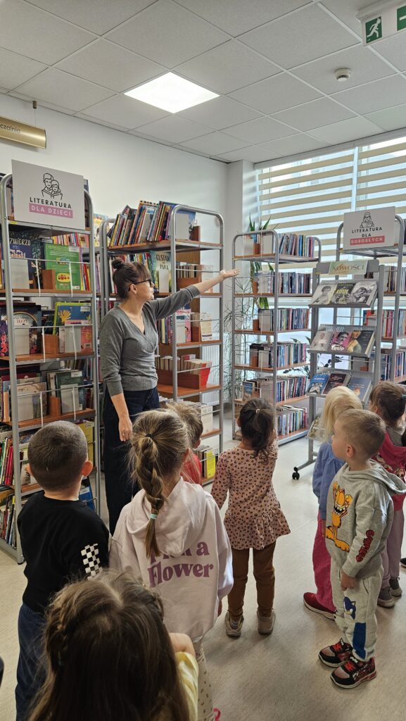 Grupa dzieci w wypożyczalni. Bibliotekarka stoi i opowiada. W tle regały z książkami.