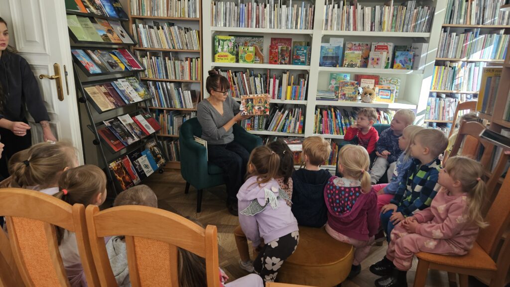 Bibliotekarka siedzi w zielonym fotelu. Przed nią siedzi troje dzieci. Z tyłu regał z książkami. Bibliotekarka pokazuje przestrzenną książkę.