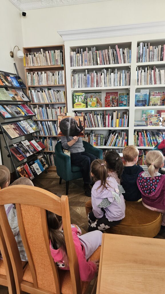 Bibliotekarka siedzi w zielonym fotelu. Przed nią siedzi grupa dzieci. Z tyłu duży regał z książkami i stojak z czasopismami. Bibliotekarka pokazuje otwartą książkę, z obrazkiem wilka z grubym brzuchem.
